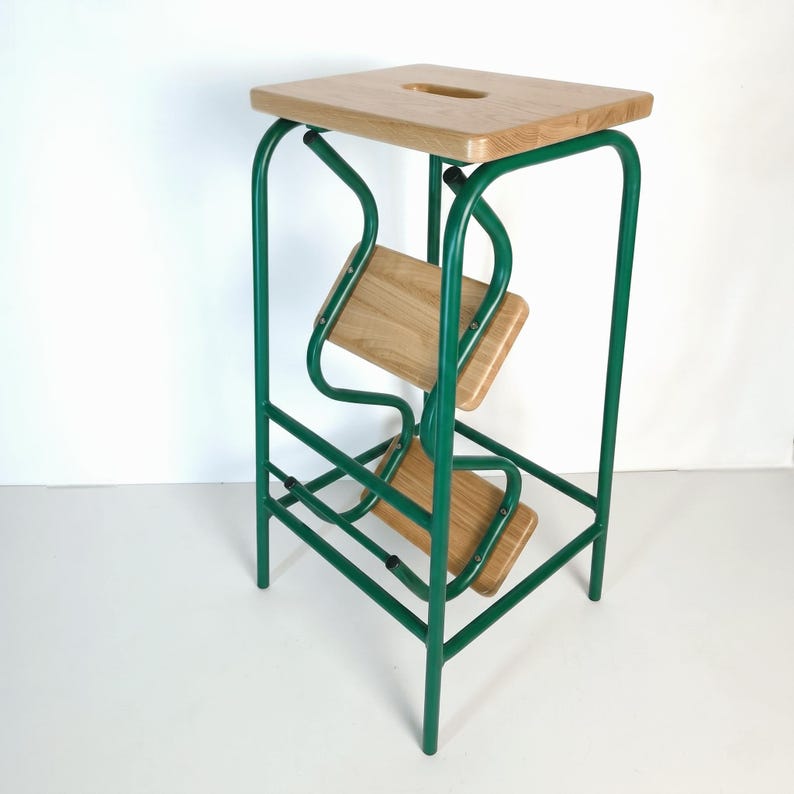 Op de afbeelding: Een groene metalen en houten opstap met een vierkante bovenkant en twee lagere treden. Het frame is heldergroen en de treden en bovenkant zijn van lichtgekleurd hout. De kruk heeft een uniek, modern ontwerp.