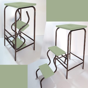 Op de afbeelding: Een unieke opstap met een saliegroene zitting en treden. Het frame is donkerbrons van kleur. De kruk heeft een vierkante bovenkant, twee treden en een kenmerkend, gebogen ontwerp. Afmetingen: ongeveer 38 cm breed, 38 cm diep en 76 cm hoog.