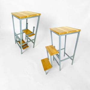 Puede incluir: Dos taburetes de bar modernos con tapas de madera clara y asientos en forma de escalón. Los marcos son de metal gris claro. Los taburetes tienen un diseño único, con un asiento pequeño y reposapiés. Sobre fondo blanco.