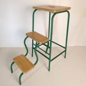 Op de afbeelding: Een vintage opstapje met drie houten treden en een bovenste platform. Het frame is gemaakt van groen metaal. Het bovenste platform heeft een rechthoekige vorm met een uitsparing. De treden zijn van licht hout.