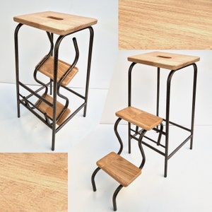 Op de afbeelding: Een houten opstapje met een rechthoekige bovenkant en twee lagere treden. Het frame is gemaakt van donker metaal met een uniek, gebogen ontwerp. Het hout heeft een lichtbruine kleur en een zichtbare nerf.
