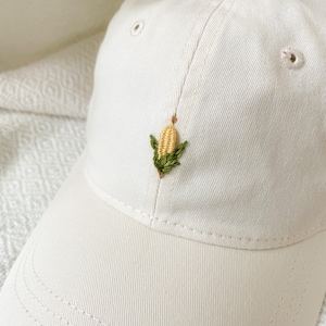 May include: Off-white baseball cap with a small, embroidered corn cob design in yellow, green, and brown. The cap has a curved brim and a classic six-panel design. The cap is made of a soft, textured fabric.