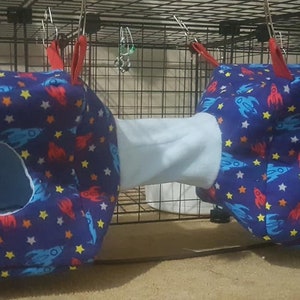May include: A navy blue pet hammock featuring a space-themed design of rockets and stars. The hammock has two cubical compartments linked by a light blue tunnel. Red straps are attached for hanging, providing a cosy space for small pets.