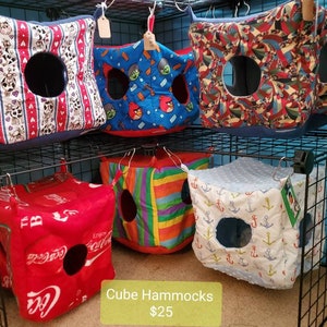 Puede incluir: Exhibición de hamacas cúbicas con diversos diseños: vacas, Angry Birds y logotipos de Coca-Cola. Cada hamaca tiene una abertura circular y está suspendida de una rejilla metálica. El texto "Cube Hammocks" es visible.