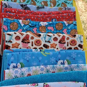 May include: A stack of colorful fabric items with various patterns, including sloths, owls, cookies, and snowflakes. The fabrics are folded and stacked on a yellow surface, showcasing a variety of designs and colors.