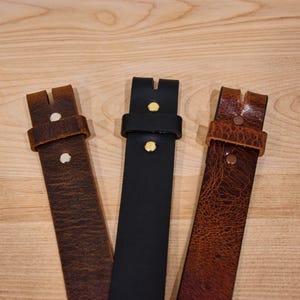 May include: Three leather belts with different colors and textures. The belts are brown, black, and a lighter brown with a textured surface. Each belt has a metal buckle with a single brass rivet.