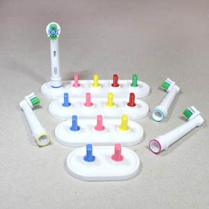 May include: White plastic toothbrush head holder with multiple colored pegs for storing electric toothbrush heads. There are four toothbrush heads in the image, two with green bristles and two with blue bristles.
