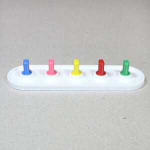 May include: A white plastic base with five colored pegs. The pegs are blue, pink, yellow, red, and green.