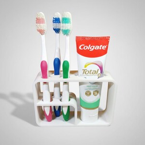 Może przedstawiać: Biały uchwyt na szczoteczki do zęb&oacute;w z trzema szczoteczkami i tubką pasty do zęb&oacute;w Colgate. Szczoteczki do zęb&oacute;w mają r&oacute;żowe, niebieskie i zielone uchwyty. Tuba pasty do zęb&oacute;w jest czerwono-biała z napisem "Colgate".