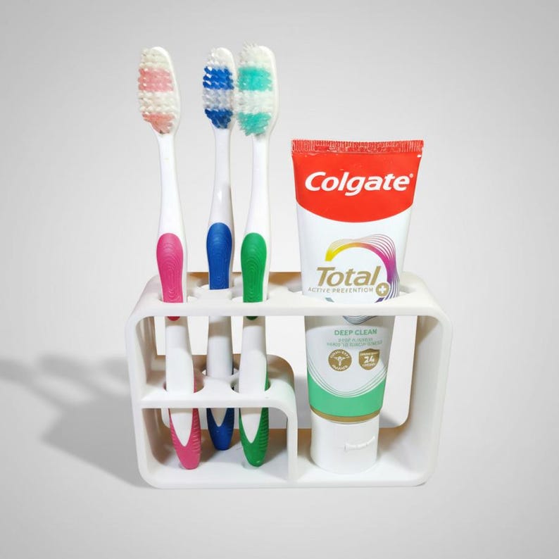 Może przedstawiać: Biały uchwyt na szczoteczki do zęb&oacute;w z trzema szczoteczkami i tubką pasty do zęb&oacute;w Colgate. Szczoteczki do zęb&oacute;w mają r&oacute;żowe, niebieskie i zielone uchwyty. Tuba pasty do zęb&oacute;w jest czerwono-biała z napisem "Colgate".