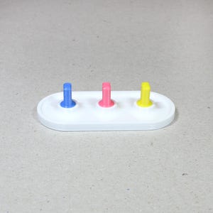 May include: A white plastic base with three colored pegs. The pegs are blue, pink, and yellow.
