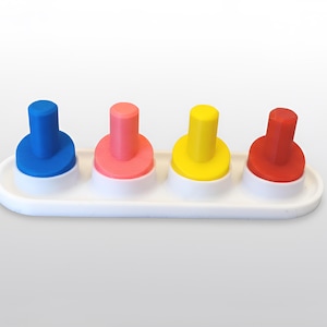 May include: A white plastic tray holds four colorful pegs: blue, pink, yellow, and red. Each peg has a cylindrical top and a circular base. The bases are white, and the tray is also white.