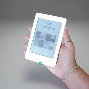 May include: A white e-reader with a green button is held in a hand. The screen displays the words "first reads" and other text. The e-reader is rectangular with rounded corners.
