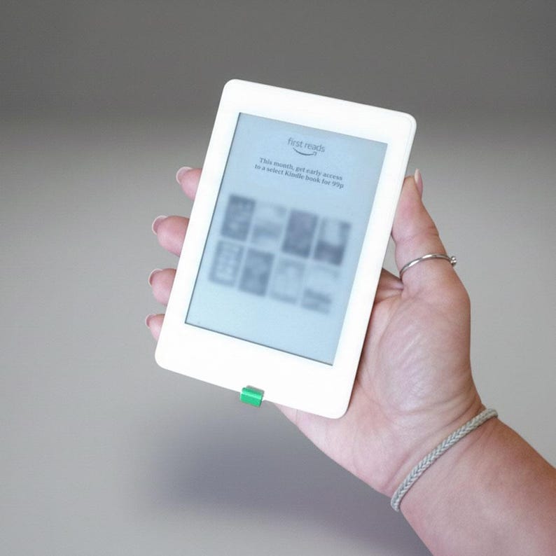 May include: A white e-reader with a green button is held in a hand. The screen displays the words "first reads" and other text. The e-reader is rectangular with rounded corners.