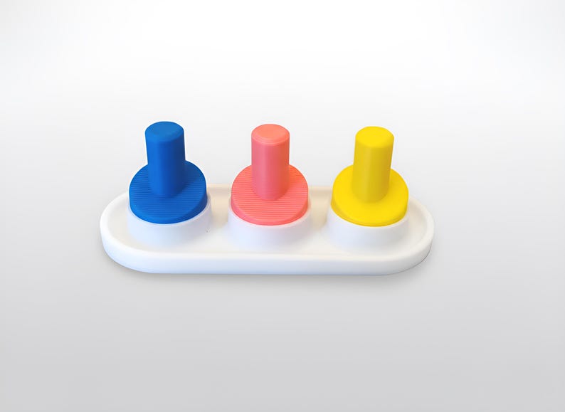 May include: A white plastic toy with three colorful pegs: blue, pink, and yellow. Each peg is cylindrical with a circular base, set on a white, oval-shaped platform. The pegs are evenly spaced and stand upright.