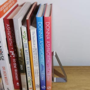 May include: A collection of books with colorful spines, including titles by Donna Ashworth, standing upright on a wooden shelf. A silver metal bookend supports the books. The books are various sizes and colors.