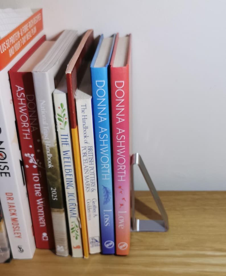 May include: A collection of books with colorful spines, including titles by Donna Ashworth, standing upright on a wooden shelf. A silver metal bookend supports the books. The books are various sizes and colors.