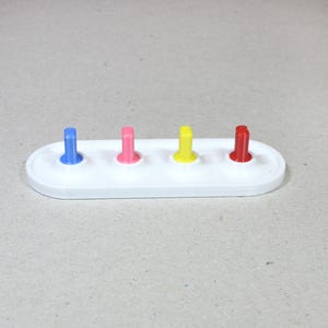 May include: A white plastic base with four colored pegs. The pegs are blue, pink, yellow, and red.