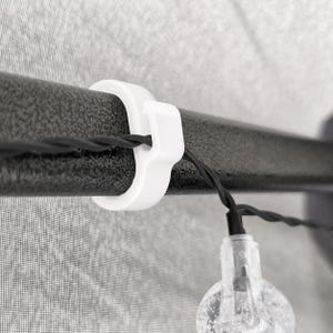 May include: A white plastic C-shaped clip attached to a textured black horizontal pole. A thin black wire runs through the clip, connecting to a clear, textured light bulb that hangs downward from the pole structure.
