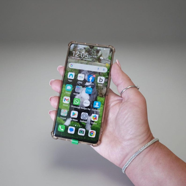 May include: A smartphone with a clear protective case is held in a hand. The phone screen displays a dog image, app icons, and the time 12:05. The case has a green accent at the bottom. A silver ring and a braided bracelet are also visible.