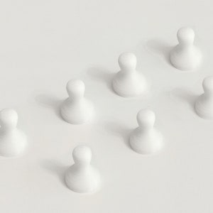 May include: Seven white, teardrop-shaped game pieces arranged in a diagonal pattern. The pieces have a smooth, rounded top and a wider base, casting soft shadows on a white surface. The overall aesthetic is minimalist and clean.