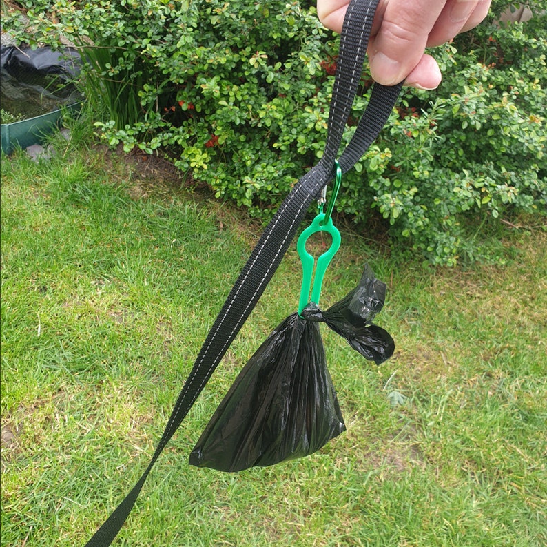 Puede incluir: Un dispensador de bolsas para excrementos de perros de pl&aacute;stico negro con un clip de pl&aacute;stico verde unido a una correa negra.