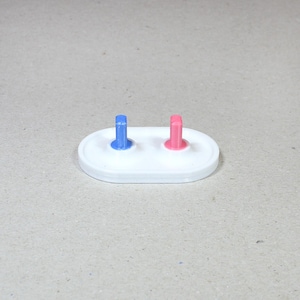 May include: A white oval base with two small, upright, colored pegs. The peg on the left is blue and the peg on the right is pink.