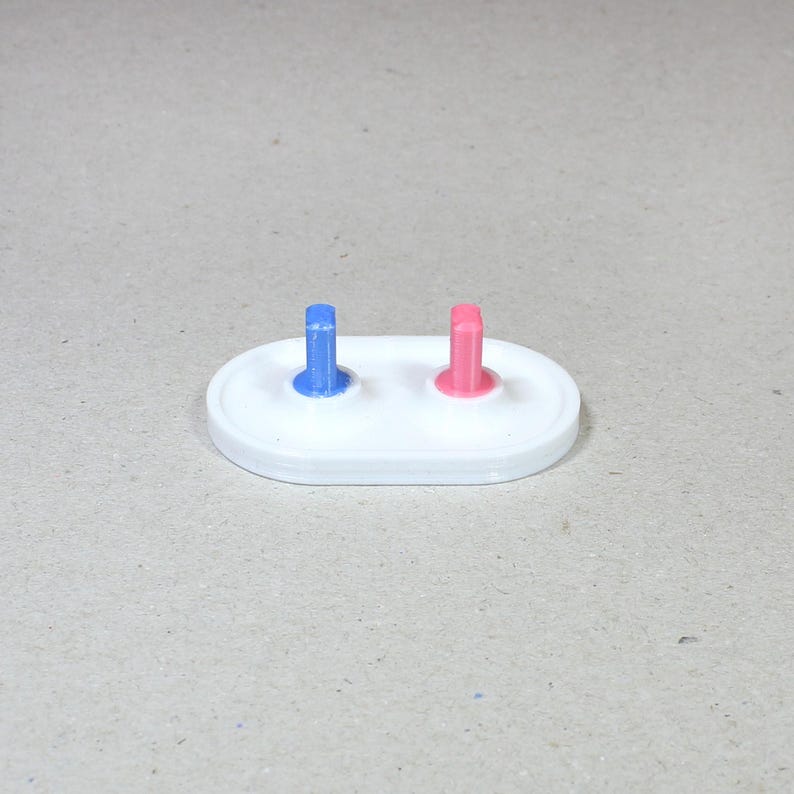 May include: A white oval base with two small, upright, colored pegs. The peg on the left is blue and the peg on the right is pink.