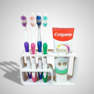 Może przedstawiać: Biały uchwyt na szczoteczki do zęb&oacute;w z czterema szczoteczkami i tubką pasty do zęb&oacute;w Colgate. Szczoteczki do zęb&oacute;w mają fioletowe, r&oacute;żowe, niebieskie i zielone uchwyty i włosie. Tuba pasty do zęb&oacute;w jest czerwono-biała ze słowem "Colgate".