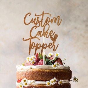 May include: A gold glitter cake topper with the words "Custom Cake Topper". The topper sits atop a layered cake decorated with strawberries and daisies. The cake has a light brown colour.