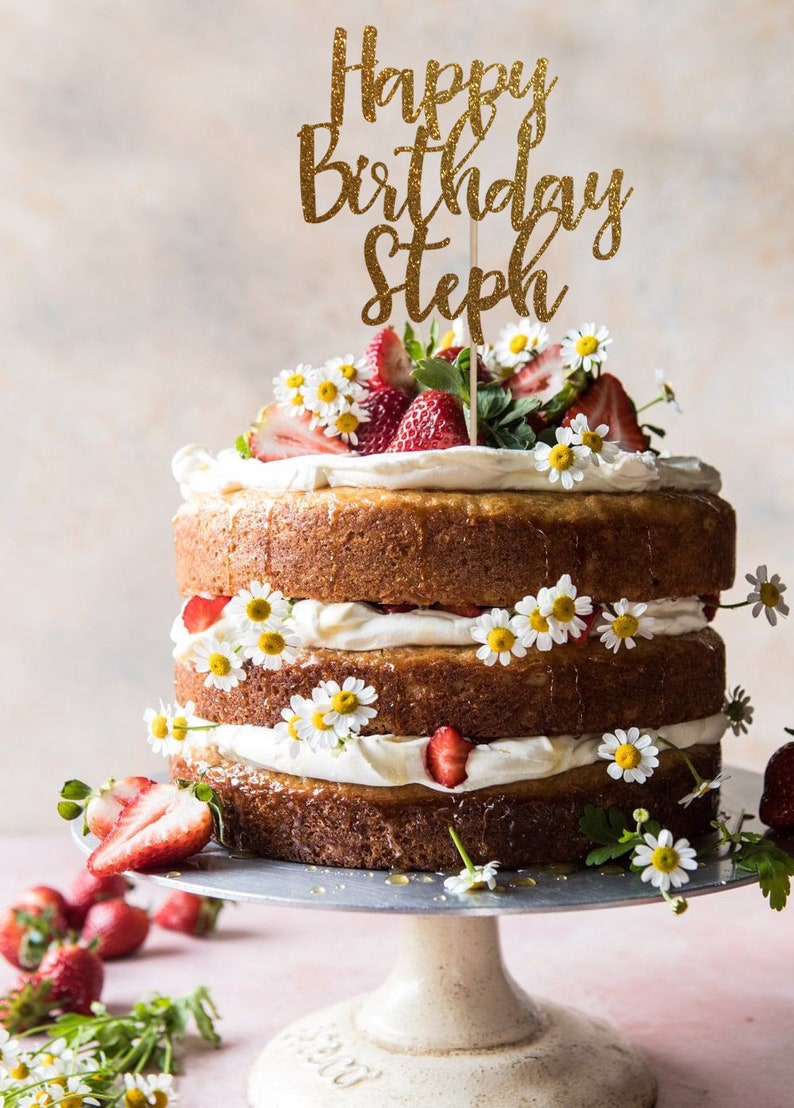 以下が含まれることがあります： イチゴとマーガレットで飾られた3段のケーキ。ケーキの上には、「Happy Birthday Steph」と書かれた金色のキラキラしたケーキトッパーが飾られています。