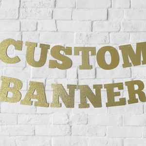 May include: Gold glitter banner with the words "CUSTOM BANNER" strung on a white brick wall.