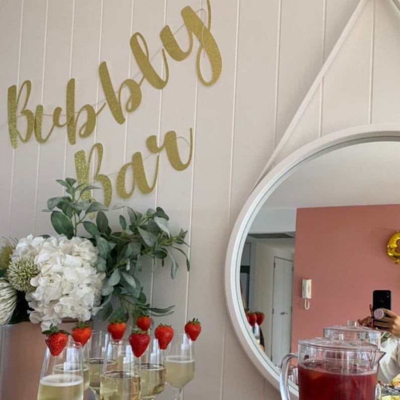 May include: A gold glitter banner reading "Bubbly Bar" is displayed on a white wall. A bouquet of white hydrangeas and greenery sits on a table in front of the banner. There are eight glasses filled with a clear liquid and topped with fresh strawberries.