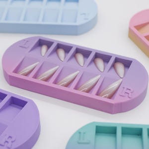May include: A collection of pastel-colored nail holders, each with a rectangular design and multiple slots. The holders are in shades of blue, purple, pink, and green. Some holders contain artificial nails. The letters "L" and "R" are printed on some of the holders.