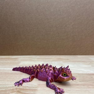 Peut inclure: Modèle imprimé en 3D d'un lézard cornu, de couleur violette et dorée. Le lézard est en position de marche, la tête tournée vers la droite. Le modèle est détaillé avec des épines et des écailles.