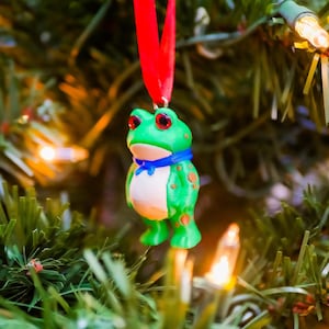 May include: A green frog Christmas ornament with red eyes and orange spots, wearing a blue scarf. The frog is suspended from a red ribbon on a Christmas tree with lights.