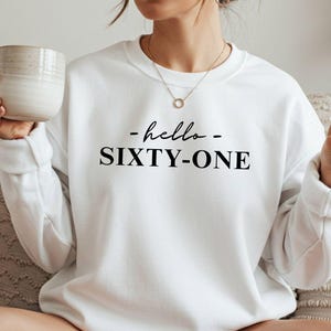 May include: White sweatshirt with the text "- hello - SIXTY-ONE" in black. The sweatshirt is a crew neck style. A person is holding a beige mug in their left hand and making a peace sign with their right hand.