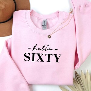 Puede incluir: Una sudadera rosa claro con el texto "- hello - SIXTY" en negro. Un collar dorado con un colgante está sobre la sudadera. Un sombrero marrón y hojas de palma secas están en el fondo.