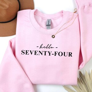 Puede incluir: Sudadera rosa claro con el texto "- hello - SEVENTY-FOUR" en negro. Un collar dorado con un colgante está sobre la sudadera. Un sombrero marrón y hojas de palma secas se ven en el fondo.