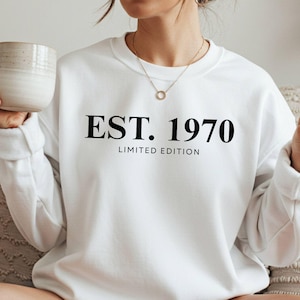 May include: White crew neck sweatshirt with the text "EST. 1970 LIMITED EDITION" in black. The sweatshirt is a solid white color. A person is holding a mug and making a peace sign.