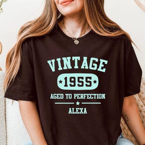 Puede incluir: Una camiseta marrón oscuro con el texto "VINTAGE 1955 AGED TO PERFECTION ALEXA" en azul claro. El texto está dispuesto verticalmente, con el año 1955 dentro de un diseño en forma de estrella.