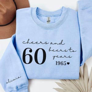 May include: Light blue sweatshirt with the text "cheers and beers to 60 years 1965". The number 60 is large and bold. The sleeve has the name "Olivia" in cursive. A brown hat is visible in the background.