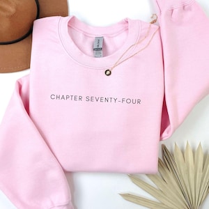 Puede incluir: Sudadera rosa claro con la frase "CHAPTER SEVENTY-FOUR" en negro. Un collar dorado con un colgante descansa sobre la sudadera. Un sombrero marrón y una hoja de palma seca también están en la imagen.