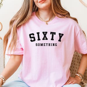 Puede incluir: Una camiseta rosa claro con las palabras "SIXTY SOMETHING" impresas en letras negras. La camiseta de manga corta parece estar hecha de un material suave. La modelo lleva la camiseta con unos vaqueros azules.