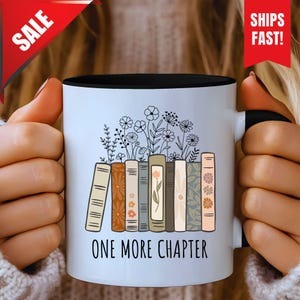 Könnte beinhalten: Eine weiße Keramiktasse mit schwarzem Innenraum, mit einem Design von Büchern mit Blumen und dem Text "ONE MORE CHAPTER". Die Bücher haben florale und gemusterte Designs. Die Tasse wird von einer Person gehalten.