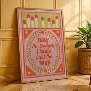 May include: A framed art print with a matchbox design. The top section shows pink match heads above white and green stripes. The centre features the text "may the bridges I burn light the way" in orange. The frame is light brown.