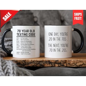May include: Two white ceramic mugs with black handles and rims. One mug reads "70 YEAR OLD TEXTING CODE" with a list of abbreviations. The other mug says "ONE DAY, YOU'RE 20 IN THE 70S. THE NEXT, YOU'RE 70 IN THE 20S."