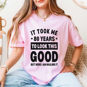 Puede incluir: Camiseta rosa claro con el texto negro: "IT TOOK ME *80 YEARS* TO LOOK THIS GOOD BUT HERE I AM NAILING IT". La camiseta de manga corta la lleva una persona. La persona lleva vaqueros azules claros.