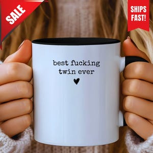 Puede incluir: Taza de cerámica blanca con interior y asa negros. La taza presenta el texto "best fucking twin ever" con un pequeño corazón negro. La taza está sujeta por una persona.