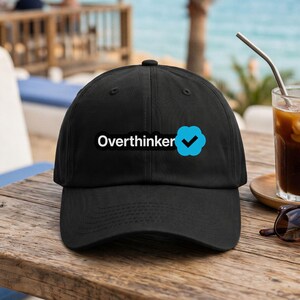 以下が含まれることがあります： 黒い野球帽で、白い文字で「Overthinker」と書かれ、その後に黒いチェックマークが入った明るい青色のバッジアイコンが付いています。この帽子は構造化されたクラウン、湾曲したつば、上部にボタンを備えた、シンプルで清潔感のあるデザインです。
